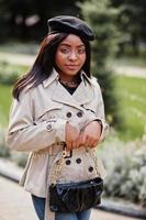 modieus Afrikaanse Amerikaans vrouw slijtage baret en jas met handtas poseren buitenshuis. foto