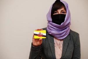 portret van jong moslim vrouw vervelend formeel dragen, beschermen gezicht masker en hijab hoofd sjaal, houden Brunei vlag kaart tegen geïsoleerd achtergrond. coronavirus land concept. foto