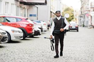 jong professioneel Afrikaanse Amerikaans videograaf Holding professioneel camera met statief pro apparatuur. afro cameraman vervelend zwart Duraq maken een videos. foto