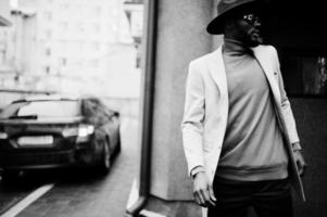 elegant Afrikaanse Amerikaans Mens slijtage beige jasje en zwart hoed met zonnebril houding in de buurt auto. foto