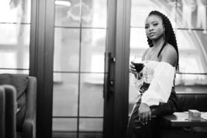 mooi Afrikaanse Amerikaans vrouw in wit blouse en rood leer broek houding Bij restaurant met glas van wijn. foto