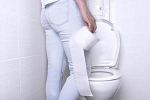 vrouw pissen, met diarree Aan een wit toilet kom in de toilet met een rollen van papier in haar hand. foto
