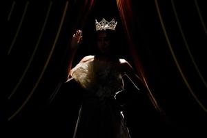 silhouet van mevrouw schoonheid optocht wedstrijd vrouw met fonkeling diamant kroon met gebruind huid mooi bedenken avond japon Aan stadium met verlichting gordijn en donker achtergrond foto