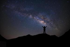 panorama landschap met melkachtig manier, nacht lucht met sterren en silhouet van een staand sportief Mens met verheven omhoog armen Aan hoog berg. foto