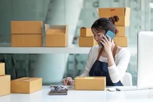 detailopname Aziatisch glimlach vrouw zijn ontvangen bestellingen via cel telefoon en Holding hand- in de buurt laptop. klein bedrijf ondernemer mkb werken met doos Bij huis, mkb e-commerce digitaal technologie concept. foto