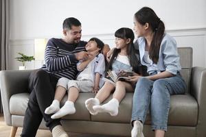 aziatische thaise familie, volwassen vader, moeder en kinderen geluk thuis wonen ontspannende activiteiten en samen een boek lezen, vrije tijd op de bank in een wit kamerhuis, heerlijk weekend, welzijn huiselijke levensstijl. foto