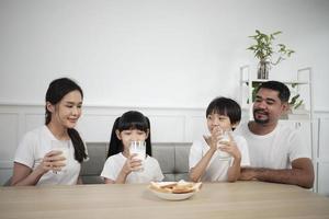 een gezond Aziatisch Thais familie, weinig kinderen, en jong ouders drinken vers wit melk in glas en brood vreugde samen Bij een dining tafel in ochtend, welzijn voeding huis ontbijt maaltijd levensstijl. foto