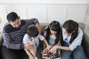 aziatische thaise familie, volwassen vader, moeder en kinderen geluk thuis wonen ontspannende activiteiten en samen een boek lezen, vrije tijd op de bank in een wit kamerhuis, heerlijk weekend, welzijn huiselijke levensstijl. foto
