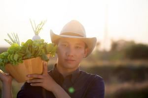 portret jonge Aziatische man glimlachend oogst en oppakken van verse biologische moestuin in mand in de hydrocultuur boerderij, landbouw en teelt voor gezonde voeding en business concept. foto