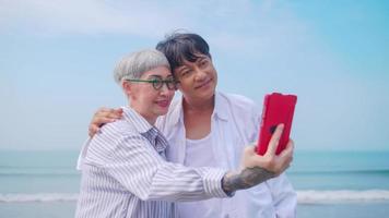 Aziatisch gepensioneerd paar ontspannende door de zee in zomer. een senior vrouw nemen een selfie foto met haar partner Aan de strand.