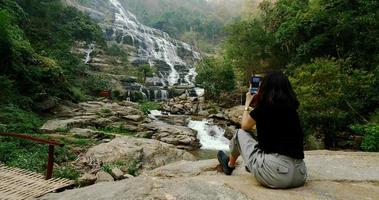 vrouw in zwart t-shirt zittend en nemen foto's van mooi waterval door smartphone. foto