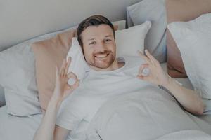 ontspannen jonge bebaarde man liggend in zijn bed en ok teken tonen foto
