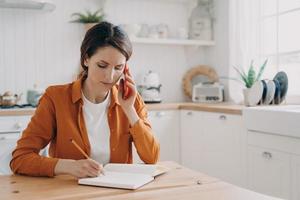 meisje zit in de keuken, maakt aantekeningen en praat aan de telefoon. officemanager afstandswerk. foto