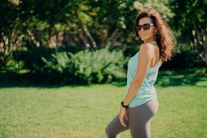 tevreden brunette europese vrouw in actieve slijtage poses zijwaarts draagt zonnebril smartwatch heeft fitness buitenshuis rust na het joggen glimlacht aangenaam. gezondheid lichaamsverzorging sport en bewegingsconcept foto