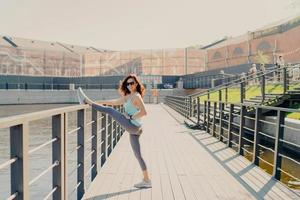 blije brunette vrouw leunt op hek rekt benen heeft perfect figuur gekleed in activewear poses buiten toont haar flexibiliteit leidt sportieve levensstijl opwarmt voor fitnesstraining. foto