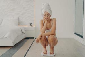 ongelukkige jonge blanke vrouw gehurkt op haar tenen over elektronische weegschalen in slaapkamer met droevige uitdrukking foto