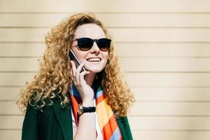 jonge lachende kantoormedewerker in zonnebril met blond krullend haar praten op mobiele telefoon tijdens een pauze staande op straat tegen beige achtergrond met kopie ruimte voor uw promotionele inhoud foto