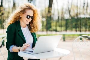 buitenshuis portret van aantrekkelijke jonge krullende vrouw zittend aan een bureau in het park met behulp van haar laptop die berichten schrijft aan haar vriend die blij is om iets aangenaams te lezen. jonge vrouw die buiten rust foto