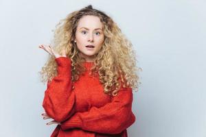 geschokt mooie blauwe ogen blonde jonge vrouw kijkt verbijsterd naar de camera, draagt losse rode trui, ontevreden met iets, geïsoleerd op witte achtergrond. verbazing begrip. foto