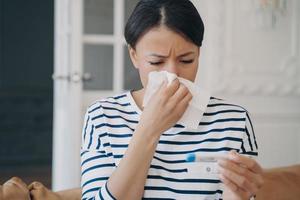 jong vrouw meten temperatuur met thermometer en blazen vloeibaar neus- in zakdoek Bij huis. foto