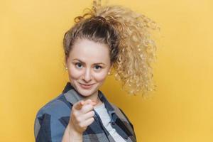 positieve zelfverzekerde mooie vrouw met krullende paardenstaart, geeft direct op camera aan, kiest jou, gekleed in casual kleding, geïsoleerd op gele achtergrond. shot van mooie vrouwengebaren binnen foto