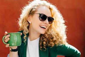 zijwaartse close-up van aantrekkelijke vrouw, gekleed in stijlvolle tinten en jas met groene kop thee met een oprechte glimlach op haar gezicht, opzij kijkend en genietend van haar leven geïsoleerd over oranje achtergrond foto