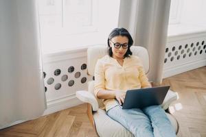 vrouw freelance werknemer werken Bij laptop zittend in fauteuil Bij huis kantoor. ver weg baan concept foto