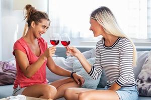twee aantrekkelijk meisjes, vrolijk het beste vrienden hebben pret en drinken rood wijn Bij huis. twee bril van wit wijn in handen foto