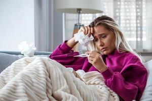 ziekte, seizoensgebonden virus probleem concept. vrouw wezen ziek hebben griep aan het liegen Aan sofa op zoek Bij temperatuur Aan thermometer. ziek vrouw aan het liegen in bed met hoog koorts. verkoudheid griep en migraine. foto