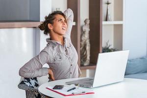 portret van jong benadrukt vrouw zittend Bij huis kantoor bureau in voorkant van laptop, aanraken pijn doen nek met gepijnigd uitdrukking, lijden van nek pijn na werken Aan pc foto