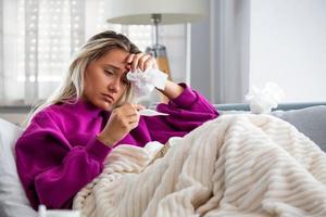 ziekte, seizoensgebonden virus probleem concept. vrouw wezen ziek hebben griep aan het liegen Aan sofa op zoek Bij temperatuur Aan thermometer. ziek vrouw aan het liegen in bed met hoog koorts. verkoudheid griep en migraine. foto