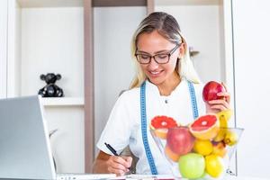 glimlachen voedingsdeskundige in haar kantoor, ze is Holding een fruit en tonen gezond groenten en fruit, gezondheidszorg en eetpatroon concept. vrouw voedingsdeskundige met fruit werken Bij haar bureau foto