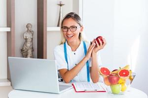 portret van jong glimlachen vrouw voedingsdeskundige in de overleg kamer. voedingsdeskundige bureau met gezond fruit, sap en meten plakband. diëtist werken Aan eetpatroon plan. foto