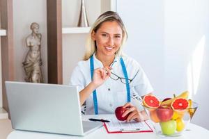 glimlachen voedingsdeskundige in haar kantoor, ze is tonen gezond groenten en fruit, gezondheidszorg en eetpatroon concept. vrouw voedingsdeskundige met fruit werken Bij haar bureau. foto