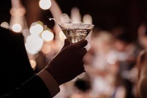 een close-up van de hand van een man die gekleed is in formele kleding en een martini-glas vasthoudt op een feestachtergrond. foto