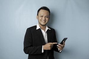 een portret van een gelukkig Aziatisch zakenman is glimlachen en Holding zijn smartphone vervelend zwart pak geïsoleerd door een blauw achtergrond foto