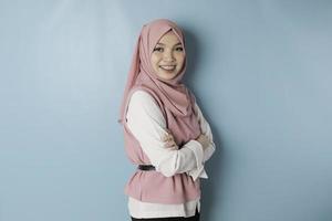 portret van een zelfverzekerd glimlachen Aziatisch moslim vrouw staand met armen gevouwen en op zoek Bij de camera geïsoleerd over- blauw achtergrond foto