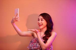 een glimlachen vrouw maken selfie foto Aan smartphone in neon licht in de studio