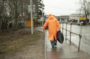 arbeider verwijdert vuilnis van weg. Mens in oranje regen. zwart zak in zijn hand. foto
