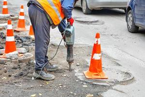 een weg arbeider werken met een elektrisch drilboor Aan een weg sectie omheind met verkeer kegels. foto