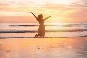 jong vrouw jumping Aan de strand Bij mooi zonsondergang, zomer vakantie concept foto