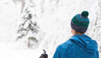 skiër in windjack en muts met pompon met skistokken in zijn handen met zijn rug tegen de achtergrond van een besneeuwd bos. langlaufen in winterbos, buitensporten, gezonde levensstijl. foto
