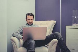 midden volwassen Mens gebruik makend van laptop terwijl ontspannende in fauteuil. foto