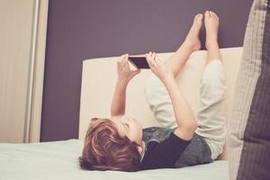 weinig jongen gebruik makend van cel telefoon en nemen selfie terwijl ontspannende Aan de bed. foto