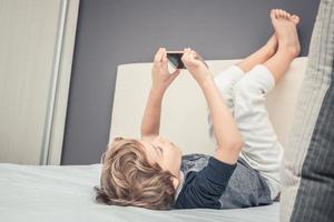 weinig jongen nemen selfie met cel telefoon terwijl aan het liegen Aan de bed. foto