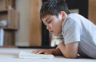 portret schoolkind leest een boek voor huiswerk, jonge jongen liggend op tapijt geniet van het lezen van het verhaal in de woonkamer, kind ontspannen thuis in het weekend. thuisonderwijs of onderwijsconcept foto