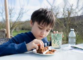 hongerig jong jongen aan het eten flapjack voor toetje, kind hebben biologisch veganistisch eigengemaakt havermout bars met veenbessen en zaden, kind hebben tussendoortje en ontspannende in de tuin Aan zonnig dag voorjaar of zomer foto