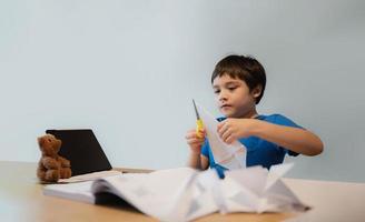 kind gebruik makend van schaar snijdend wit papier, schooljongen maken origami zwaan voor zijn huiswerk, kind aan het leren papier kunst origami les en kunst en ambacht, kinderen leren en Speel Bij huis, terug naar school- concept foto