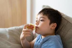 gezond kind dat rode appel eet, schattige jongen die vers fruit eet voor zijn snack terwijl hij tv kijkt in de woonkamer, close-up kind gezicht etend. gezond voedsel voor kinderen concept foto