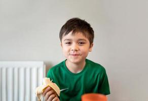 healhty jong jongen aan het eten banaan, gelukkig kind hebben ontbijt, schattig kind op zoek Bij camera met glimlachen gezicht. foto